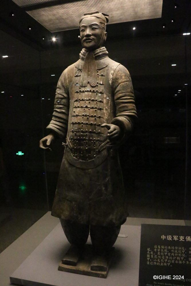 This photo shows the statue of the High-ranking Officer. This is one of seven ’generals’ found in the terra-cotta pits. The height, clothing and headgear of this officer all indicate his high rank. He wears double-layered robes under a colorful fish-scaled armor, and a high headgear tied with ribbons under the chin. His shoes have square opening and upward-bending tips. There are eight knots made of ribbons to decorate the armor, three knots on the front plate, three on the back and one knot each on the shoulder.