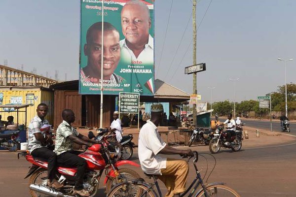 People ride bicycles and motorcycles past a campaign poster of Ghanaian President and National Democratic Congress (NDC) presidential candidate John Mahama in Tamale on December 6, 2016, eve of Ghana's presidential and parliamentary elections.